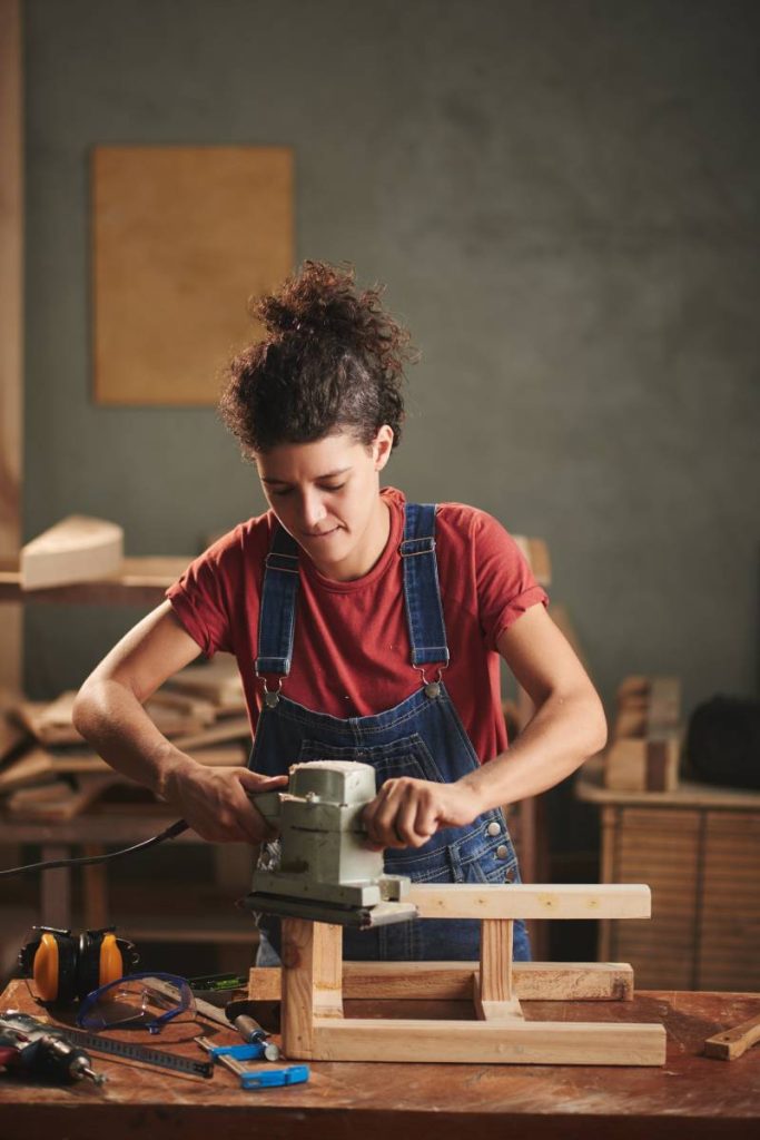 Young curly woman in denim overall enjoying process of smoothing wooden stool surface with electrical belt sander